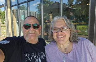 A man with sunglasses and a black shirt and a woman with glasses and a purple shirt smiling in a selfie