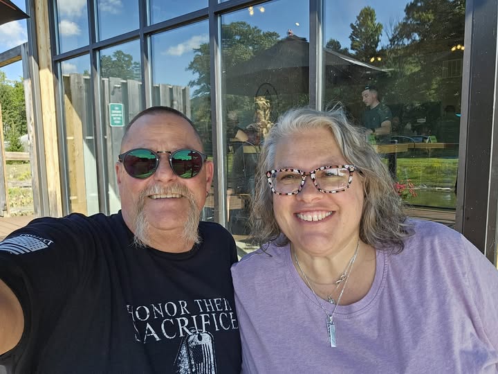 A man with sunglasses and a black shirt and a woman with glasses and a purple shirt smiling in a selfie