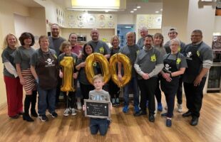 Group of people surrounding the golden balloon numbers 100, child in front holding blackboard that reads "Friends of Bear, 100th Meal Served"