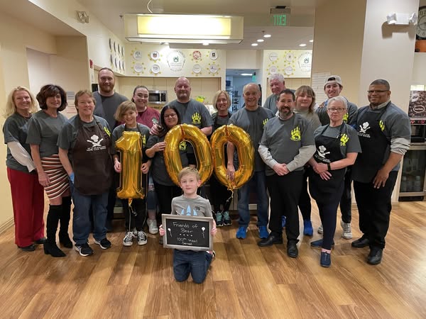 Group of people surrounding the golden balloon numbers 100, child in front holding blackboard that reads "Friends of Bear, 100th Meal Served"