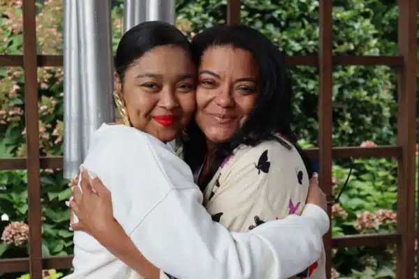 Photo depicts on the left Chelsea a lung transplant recipient smiling and hugging her smiling Mother Nicole, her caregiver. Located in front of the Gift of Life Howie's House chimes.
