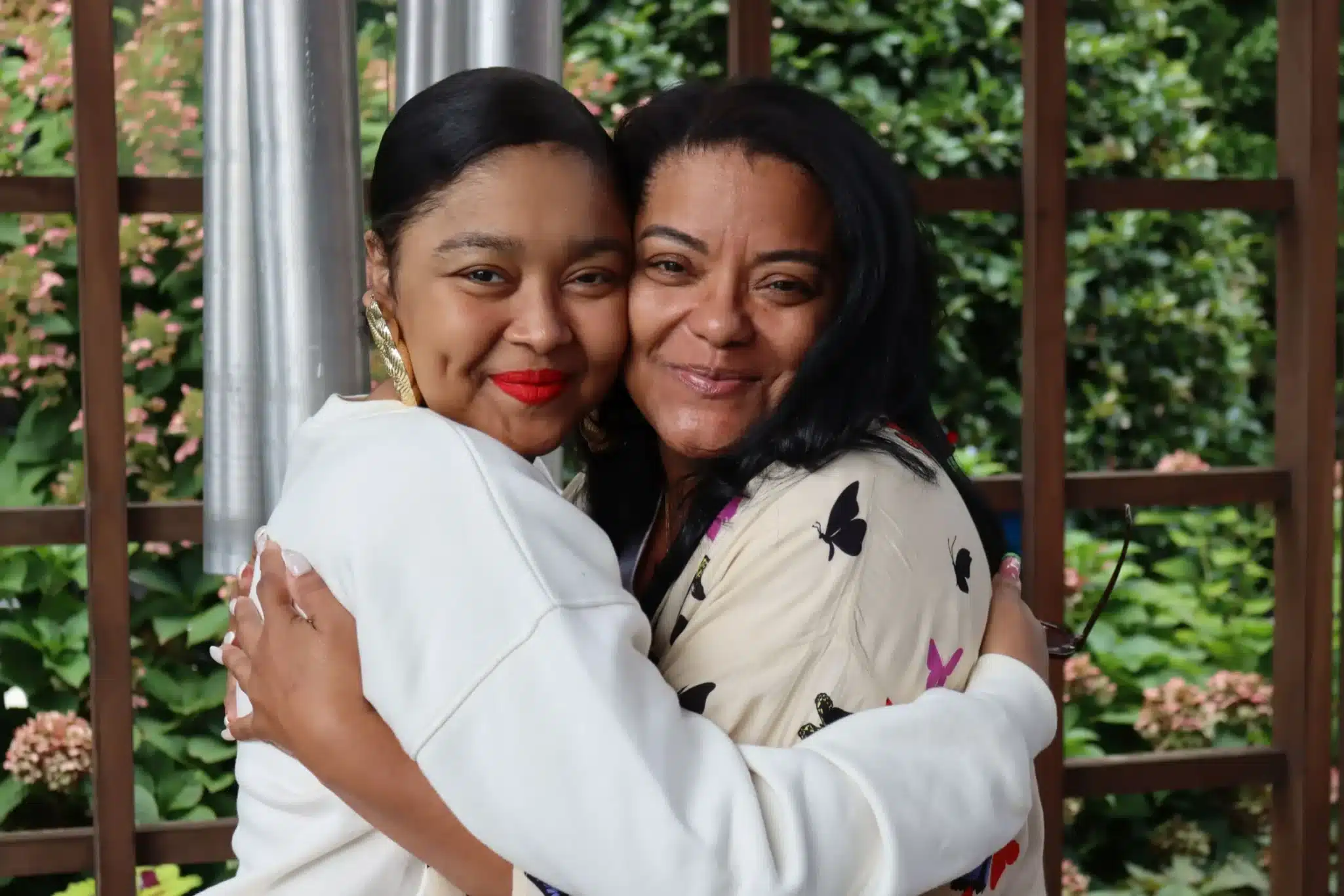 Photo depicts on the left Chelsea a lung transplant recipient smiling and hugging her smiling Mother Nicole, her caregiver. Located in front of the Gift of Life Howie's House chimes.