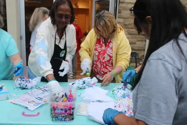 Depicts 3 women standing around a table of rubber banded t-shits squeezing bottles of dye over them