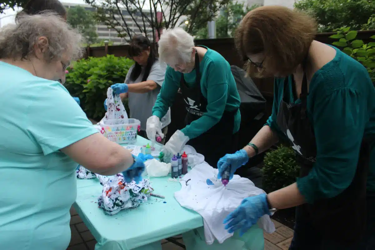 Depicts 3 women standing around a table of rubber banded t-shits squeezing bottles of dye over them