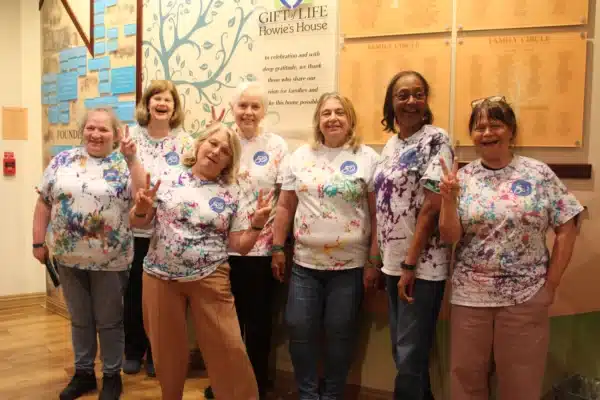 Image depicts 7 women standing side by side in Gift of Life Howie's House Foyer showing off their freshly made tie dye shirts