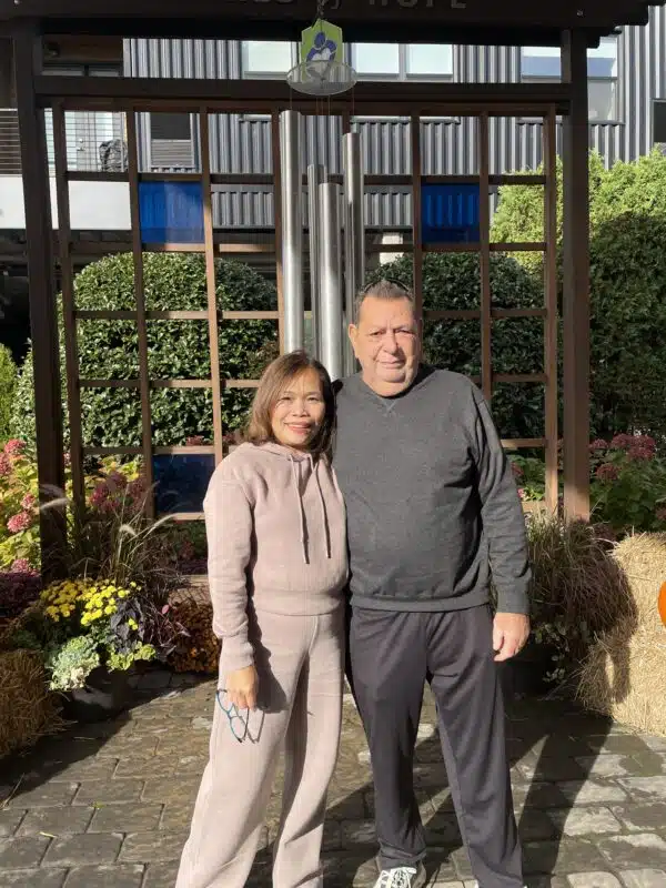 A light brown haired Asian woman in a beige sweat set stands with her arm around a taller Caucasian man in a black long sleeved shirt and black pants. They are standing in from of a large hanging chime with flowers and haybales in the background,