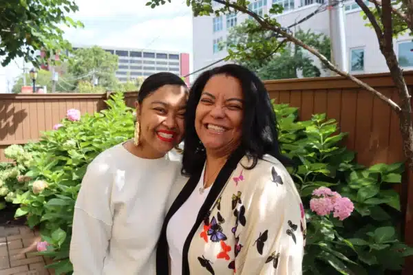Photo depicts on the left Chelsea a lung transplant recipient smiling and hugging her smiling Mother Nicole, her caregiver. Located in front of the Gift of Life Howie's House chimes.