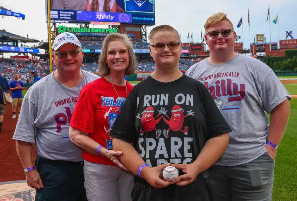 A blonde family of four stands in a baseball stadium. They are wearing Philadelphia Phillies merchandise. A young man in the middle is wearing a shirt that has "I run on spare parts" with a graphic of two kidneys, he is holding a baseball.